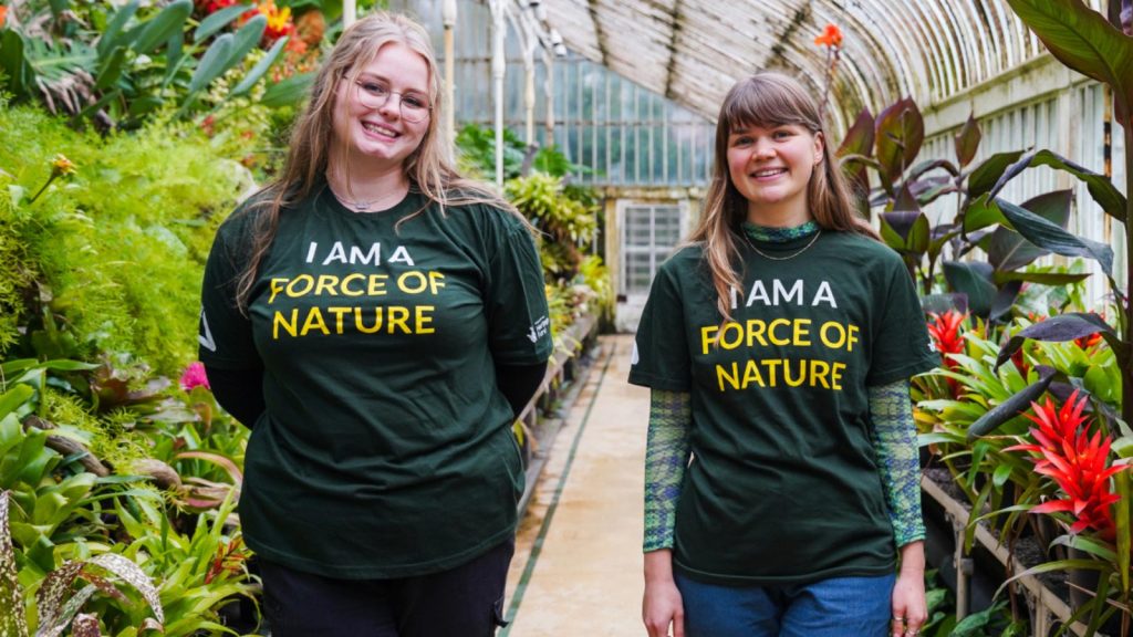 Two young women standing in a greenhouse. Both wearing green T Shirts with the words "I am a force of nature".