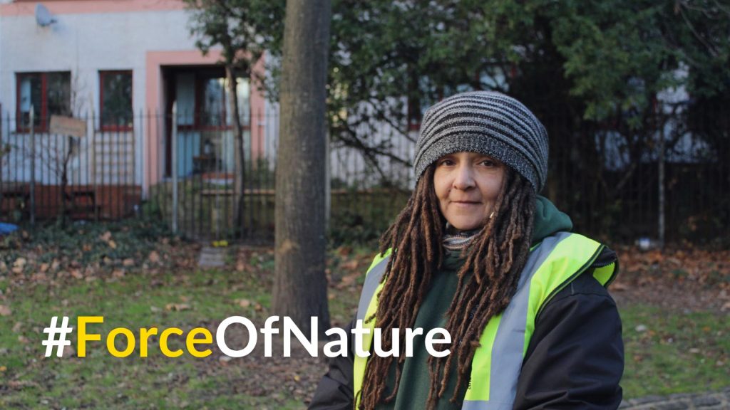 A woman with plaits and a beanie hat standing in a garden.